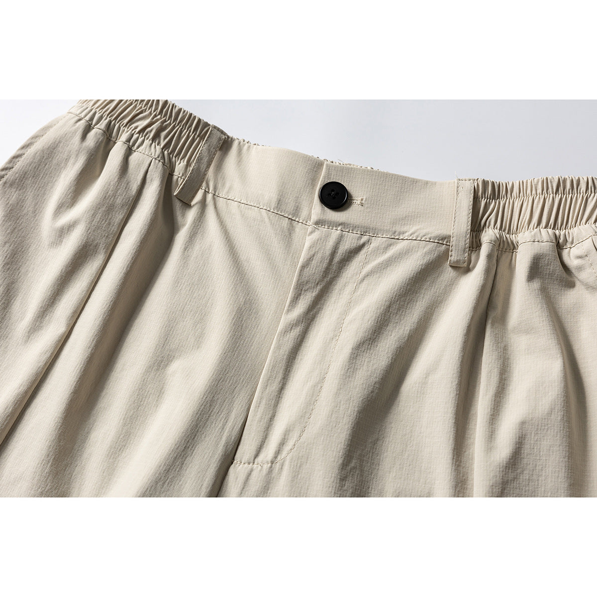 Close-up of cream trousers showing the waistband, black button, belt loops, elasticated sides and front pleats.