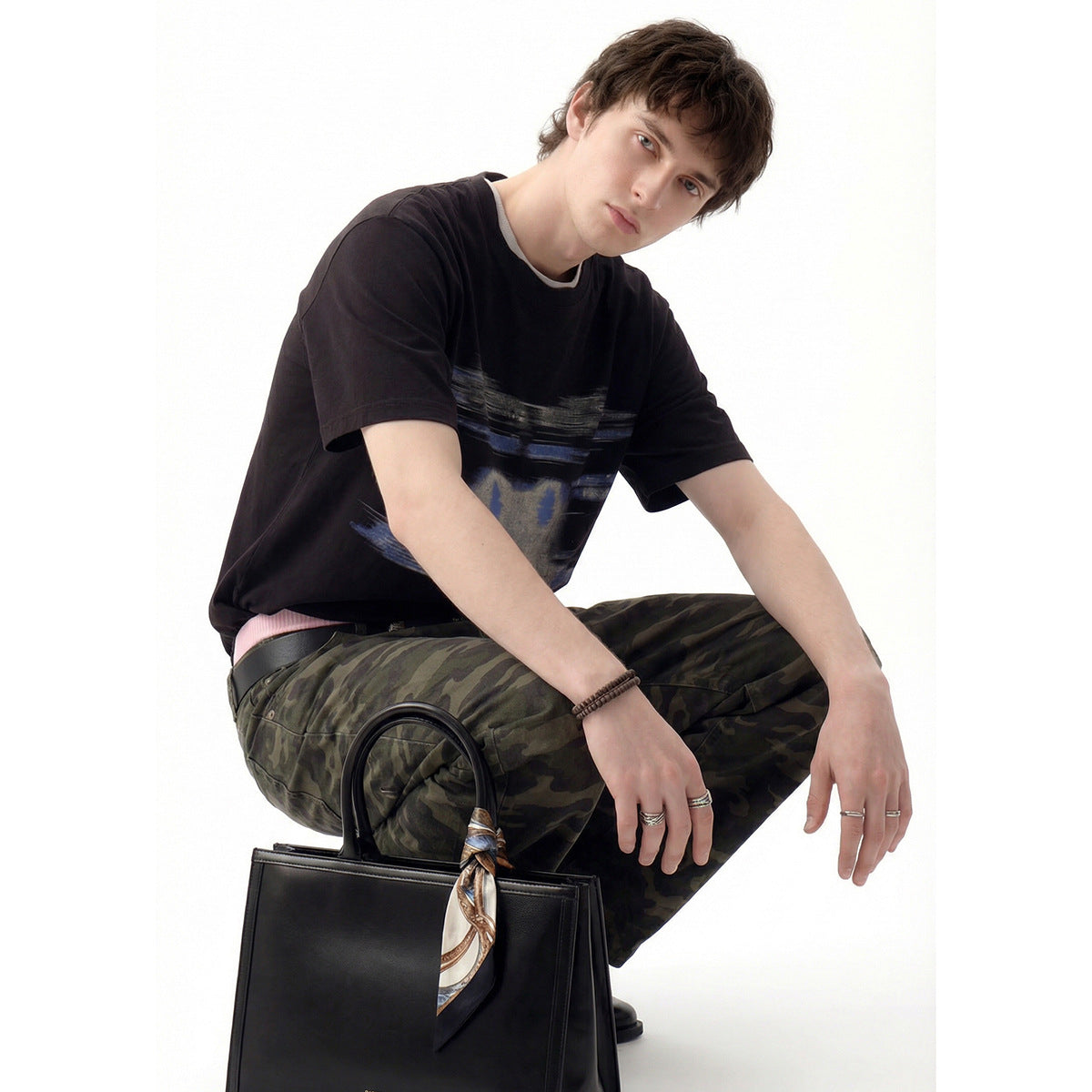 Model crouching in a black short-sleeve T-shirt with an abstract blue-gray chest graphic and camouflage pants.