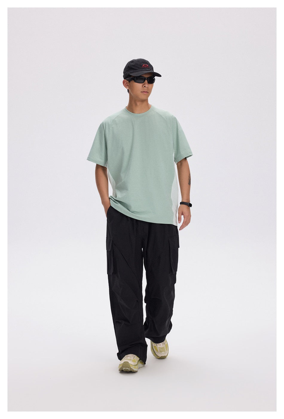Model wearing a pale mint oversized T-shirt and loose black cargo pants, with a black cap and sunglasses.