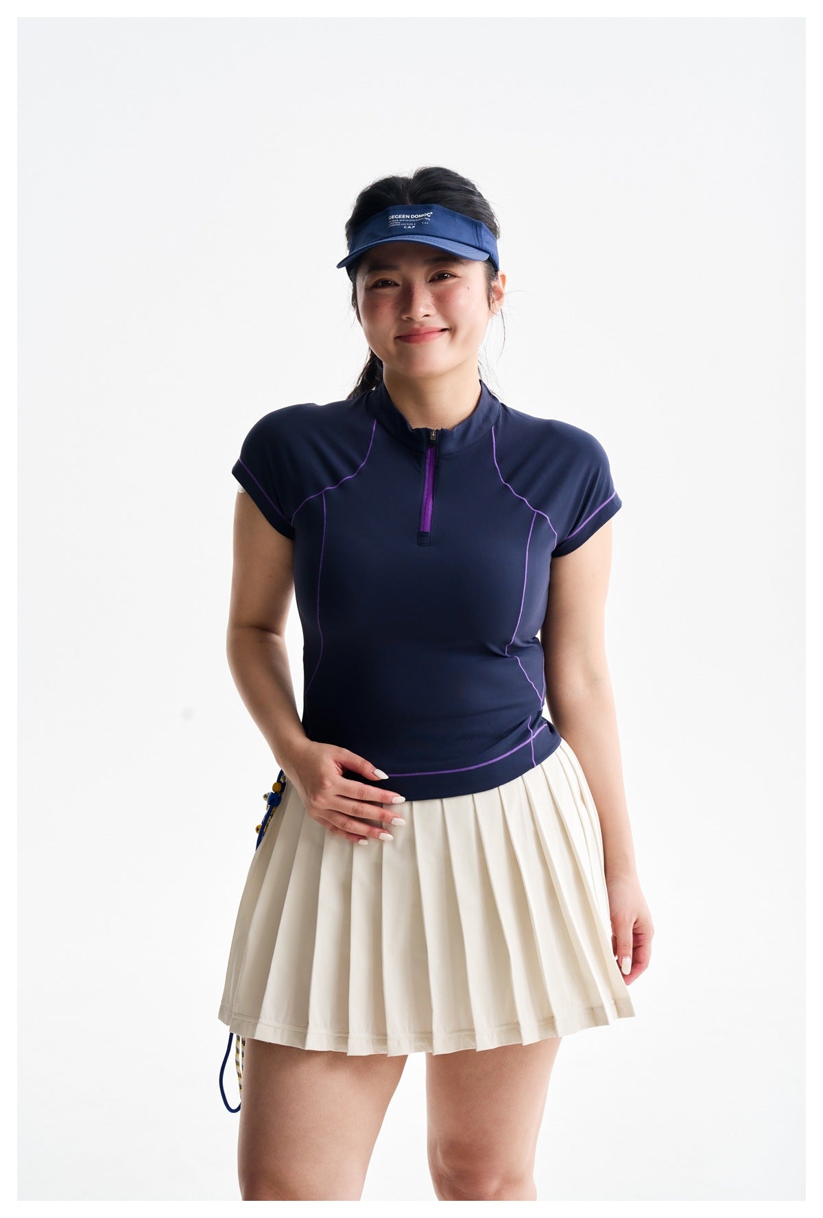 Model wearing a navy cap-sleeve zip-neck top with purple seam accents and a cream pleated tennis skirt.