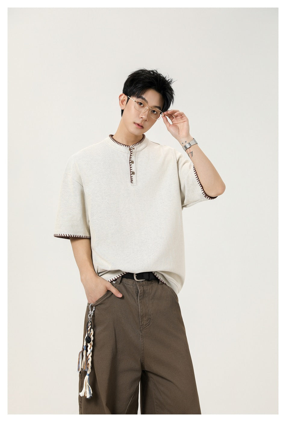 Model wearing an ivory short-sleeve henley with dark contrast stitching and brown trousers.