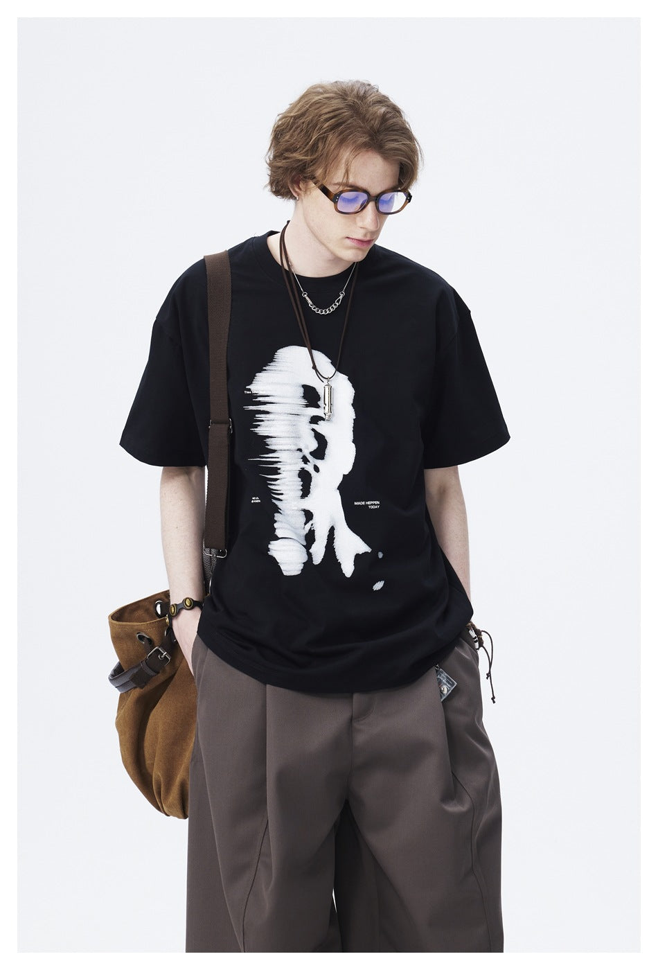 Model wearing a black T-shirt with a large white abstract face graphic, shown with loose dark trousers.