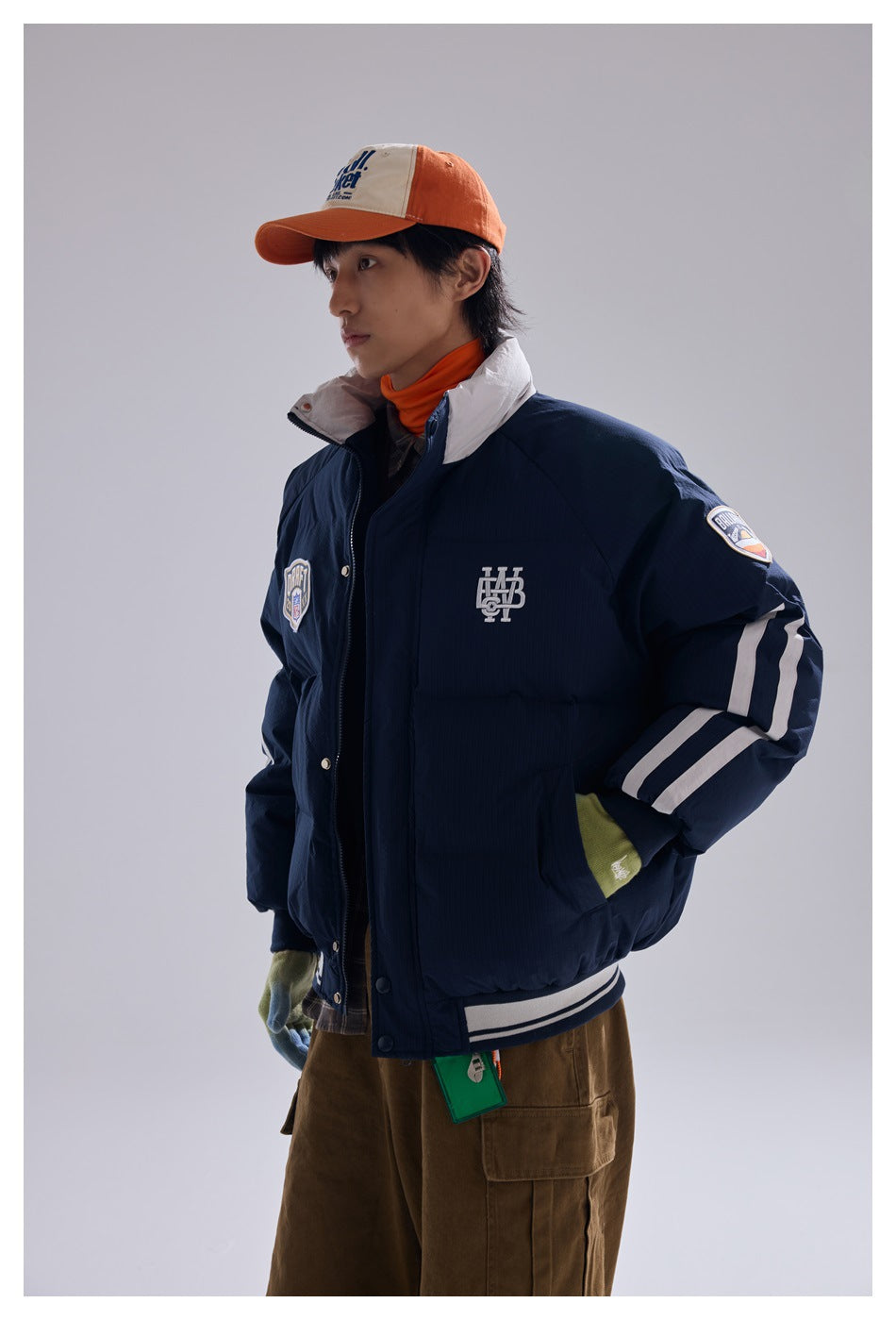 Model wearing a navy puffer jacket with a white collar, sleeve stripes and patches, shown from the front at an angle.