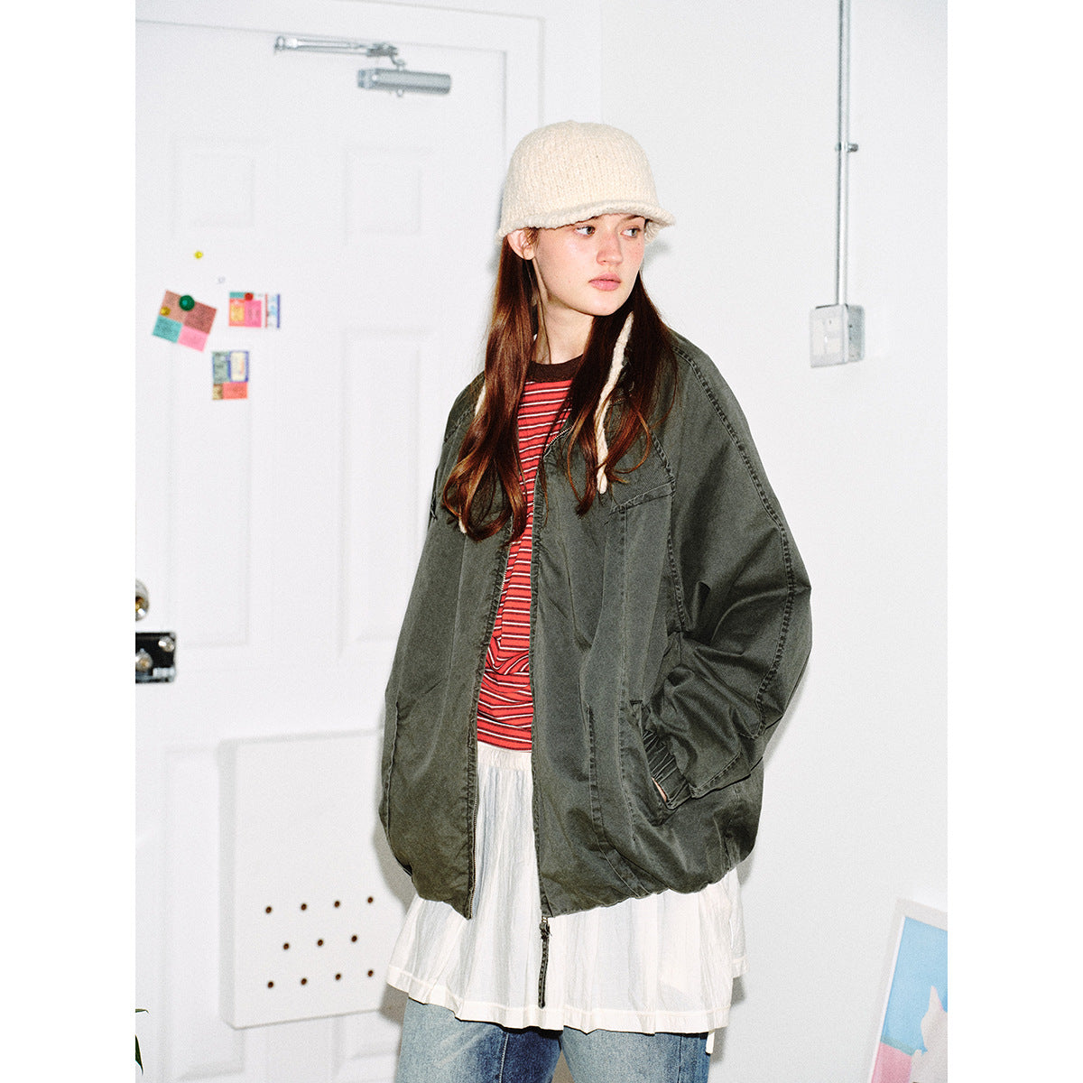 Model wearing an oversized dark olive jacket over a red striped top and white skirt, with a cream knit hat.
