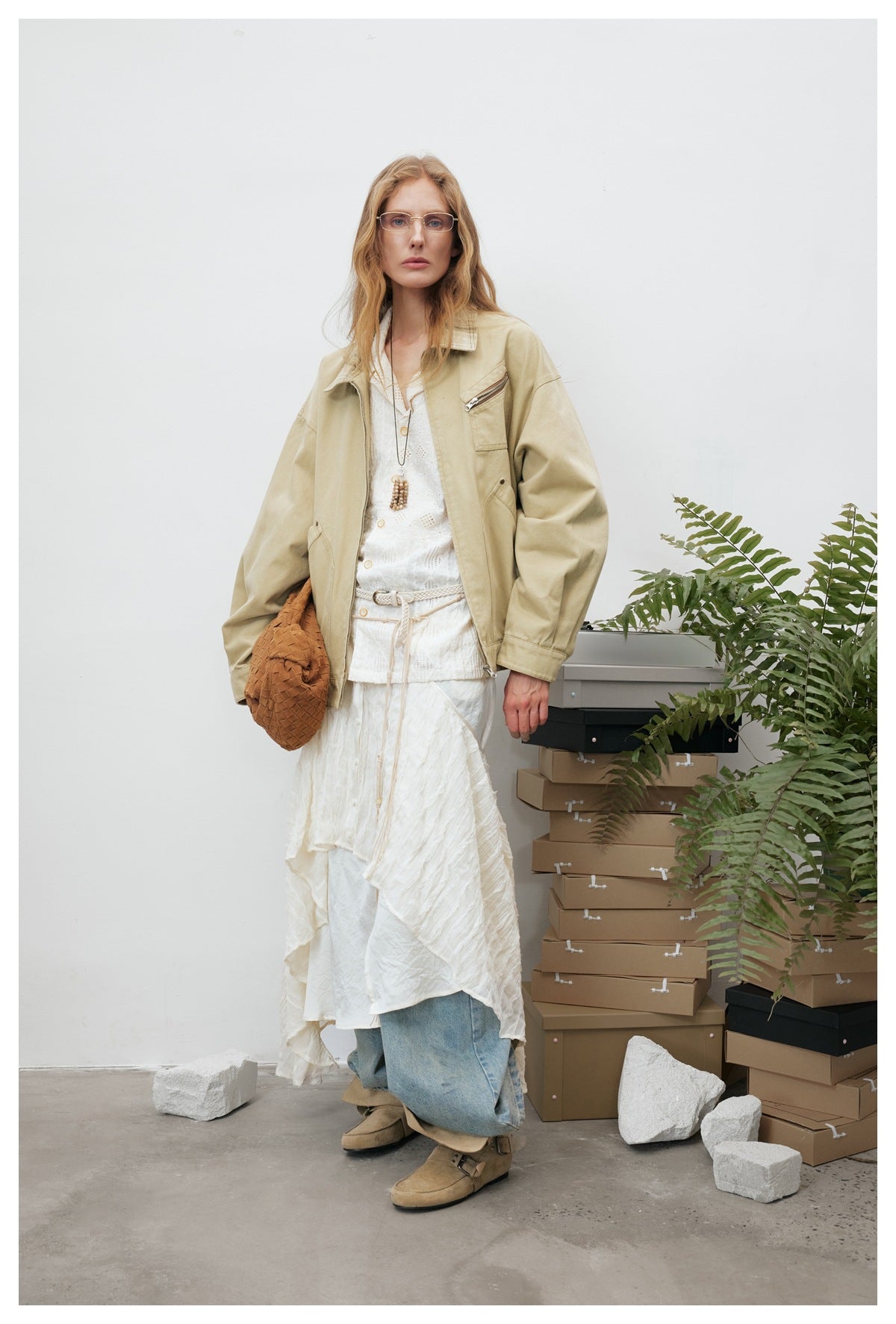 Model wearing an oversized khaki zip jacket over a white shirt and long skirt with jeans, standing by a plant