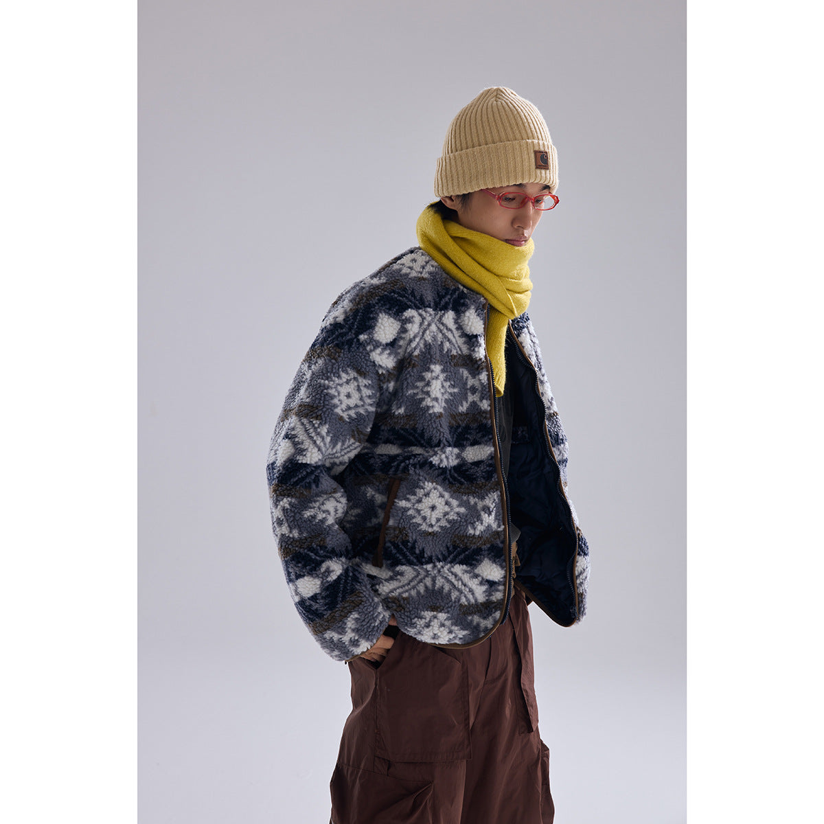 Model wearing a gray, navy, and cream geometric fleece jacket with a yellow scarf and beige beanie, shown at an angle.