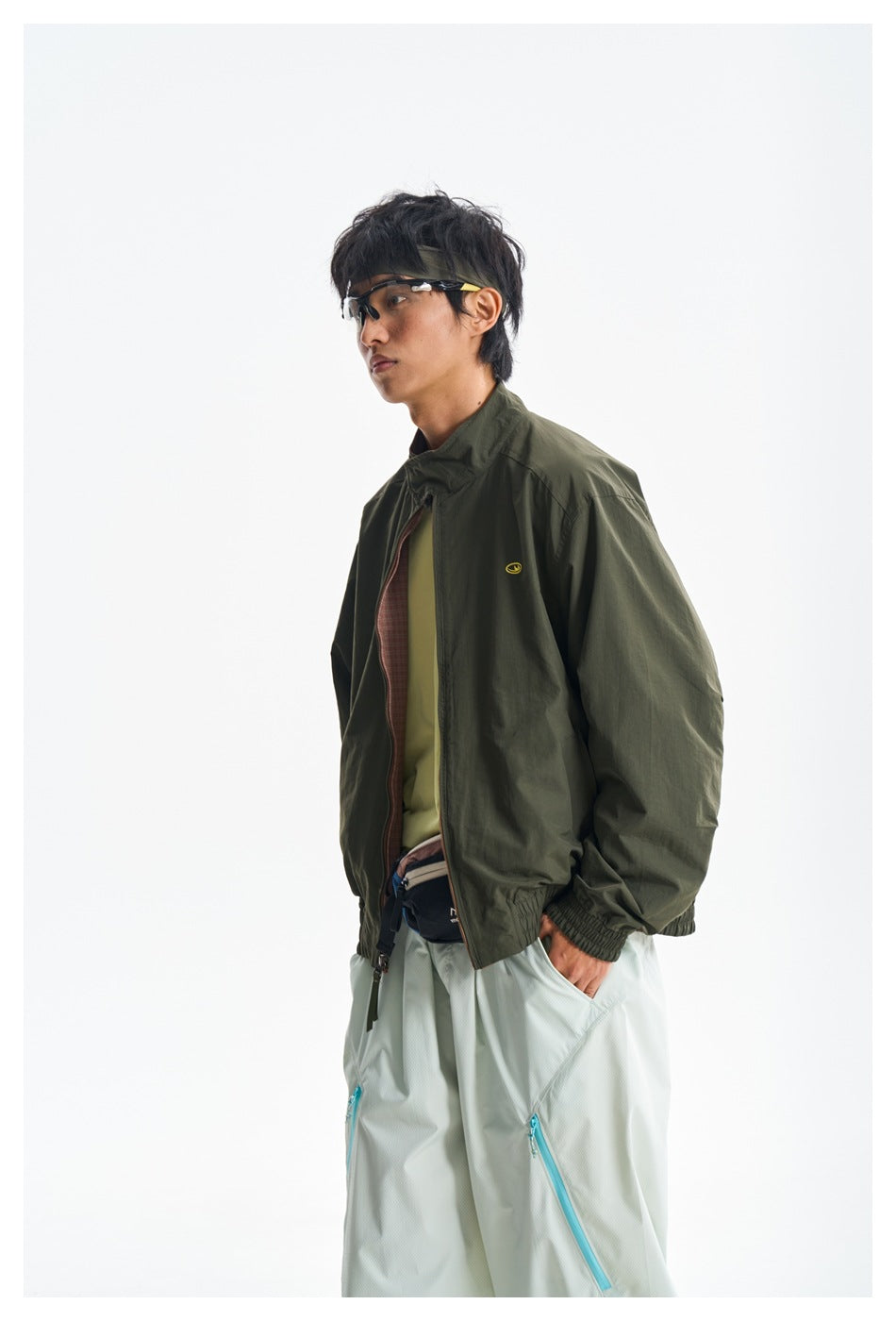 Closer view of a model in a dark olive jacket and pale yellow shirt with loose white trousers and turquoise zippers.