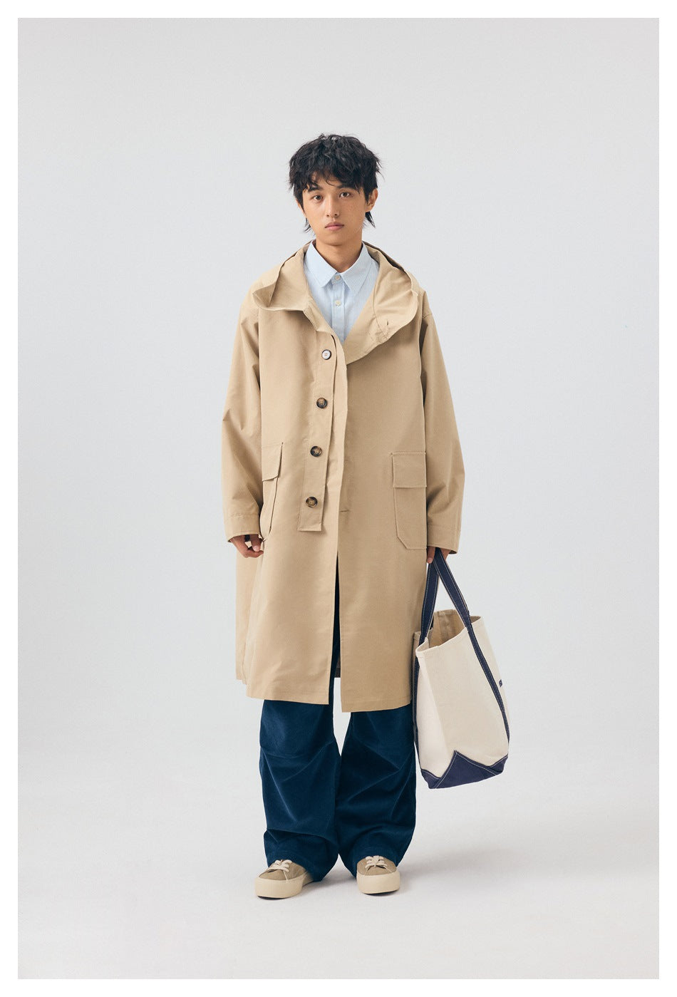 Model wearing a khaki long hooded coat over a striped shirt with navy pants, holding a canvas tote
