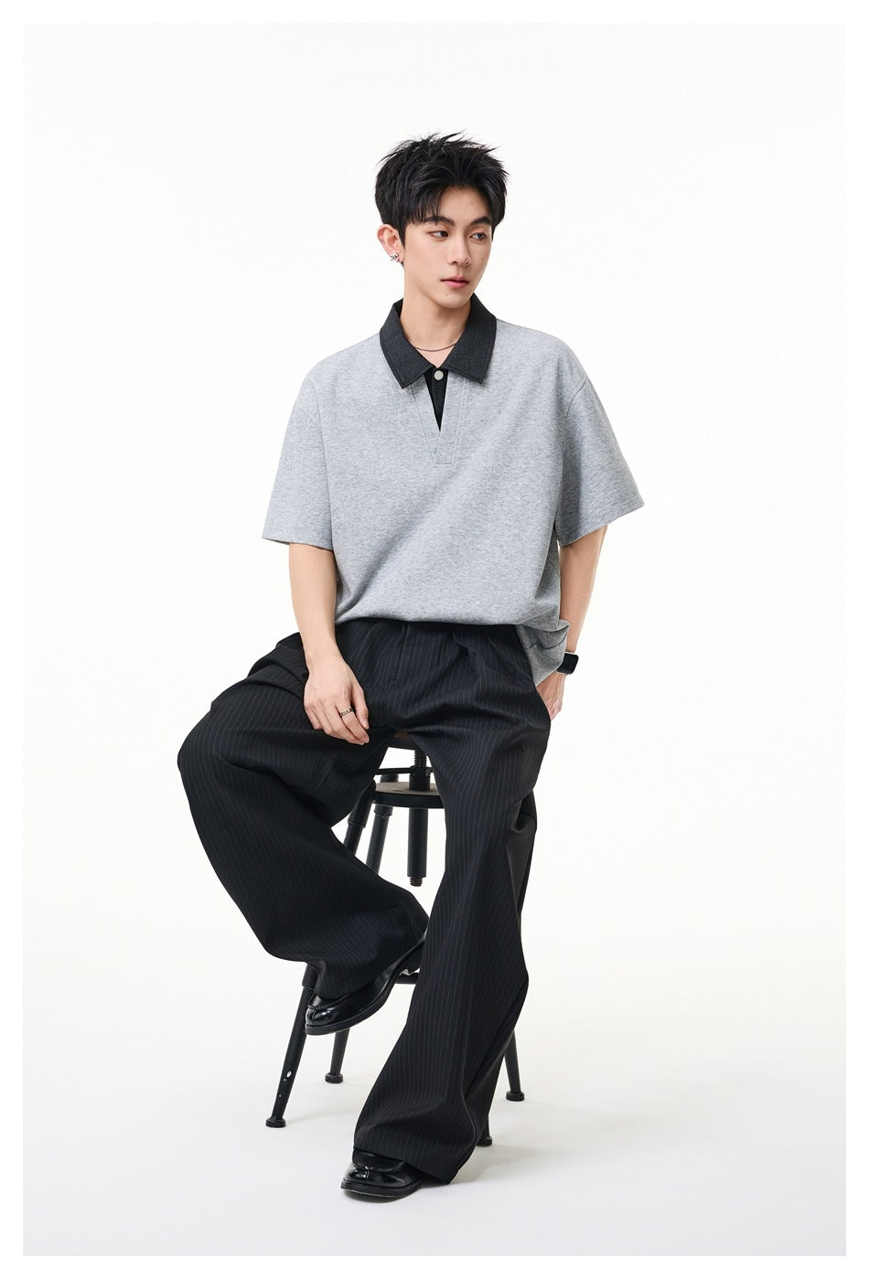 Model seated in a gray short-sleeve polo with a dark collar and black pinstripe trousers.