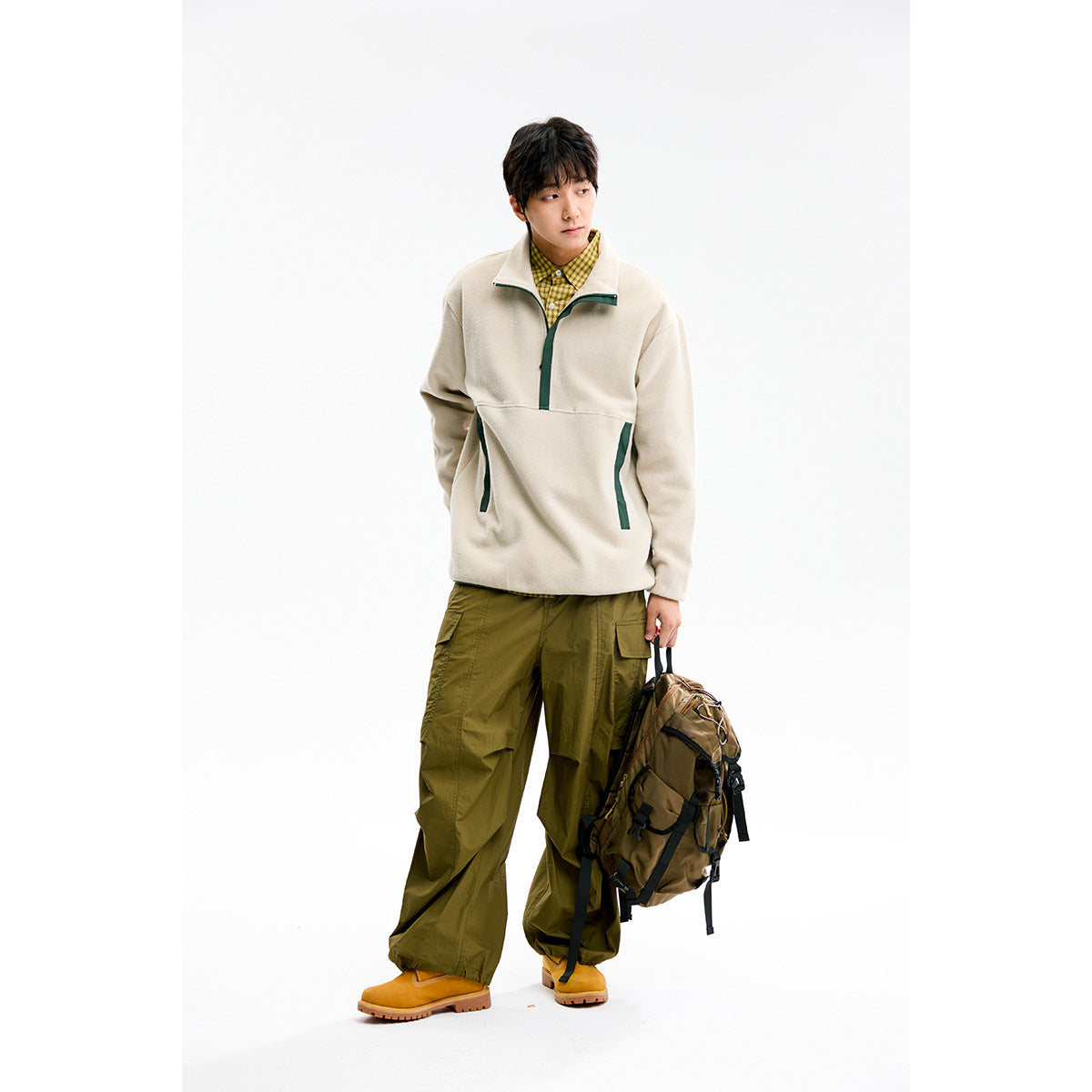 Model wearing a cream pullover with dark green trim, a yellow checked shirt, olive cargo pants, and carrying a backpack.