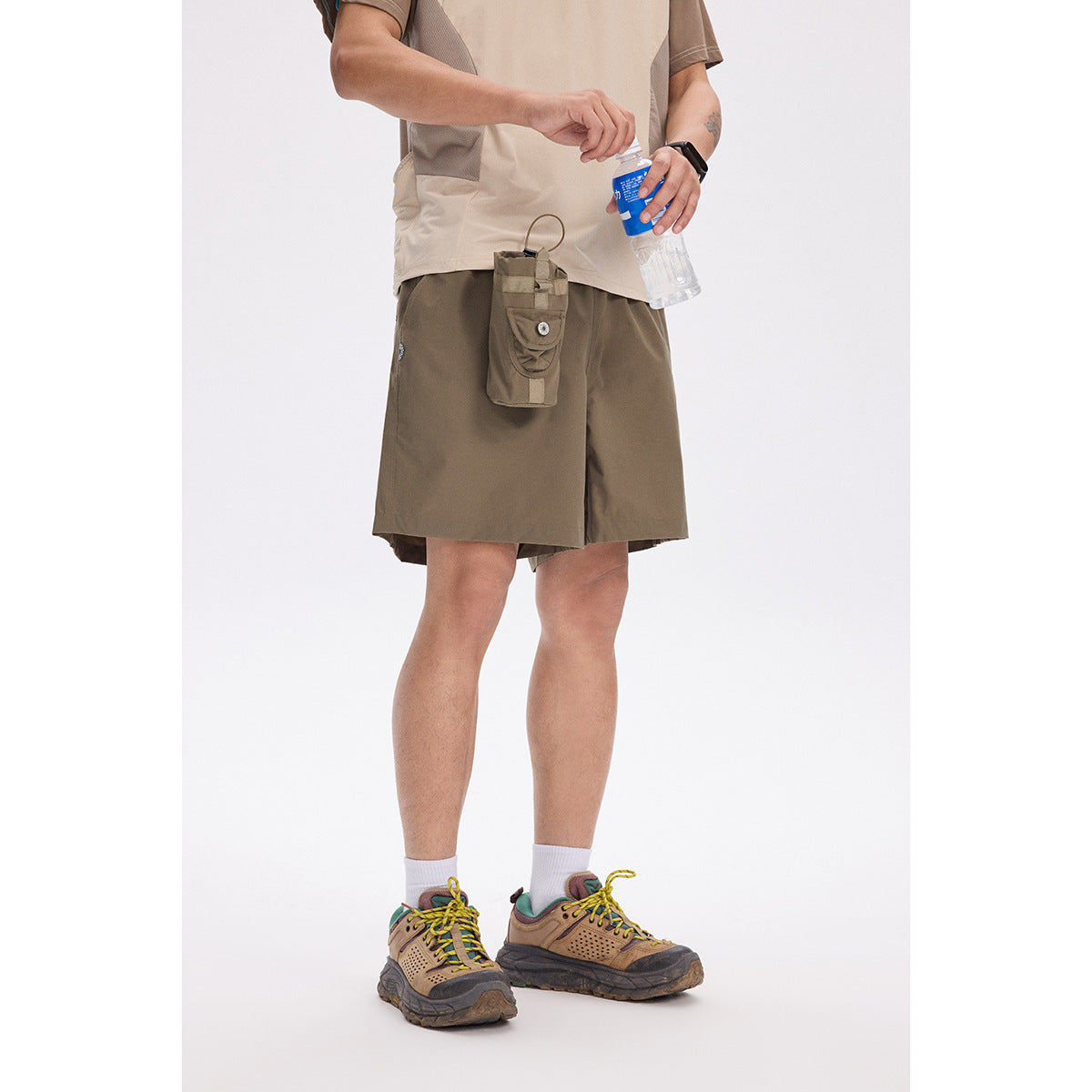 Person wearing olive shorts with a hanging utility pouch, beige shirt, white socks, and trail shoes.