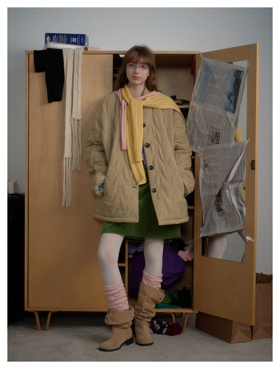 Model wears a tan wave-quilted button-front coat over a green dress, with a yellow scarf and pink accents.