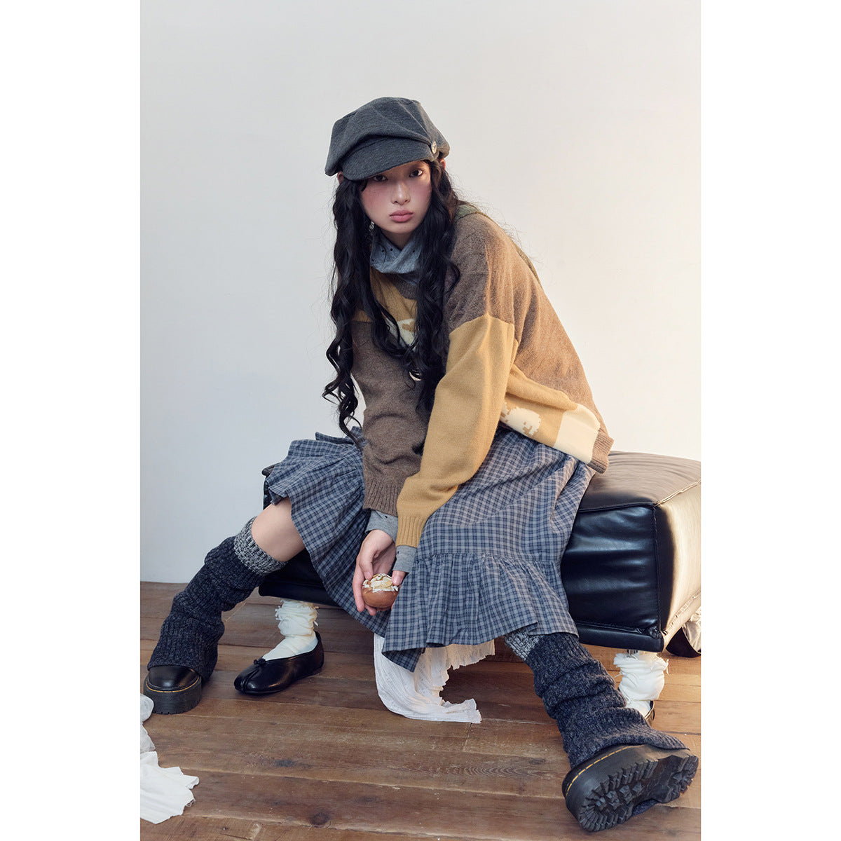 Model seated in a brown and mustard cardigan and blue plaid tiered skirt, with dark leg warmers and a gray cap.