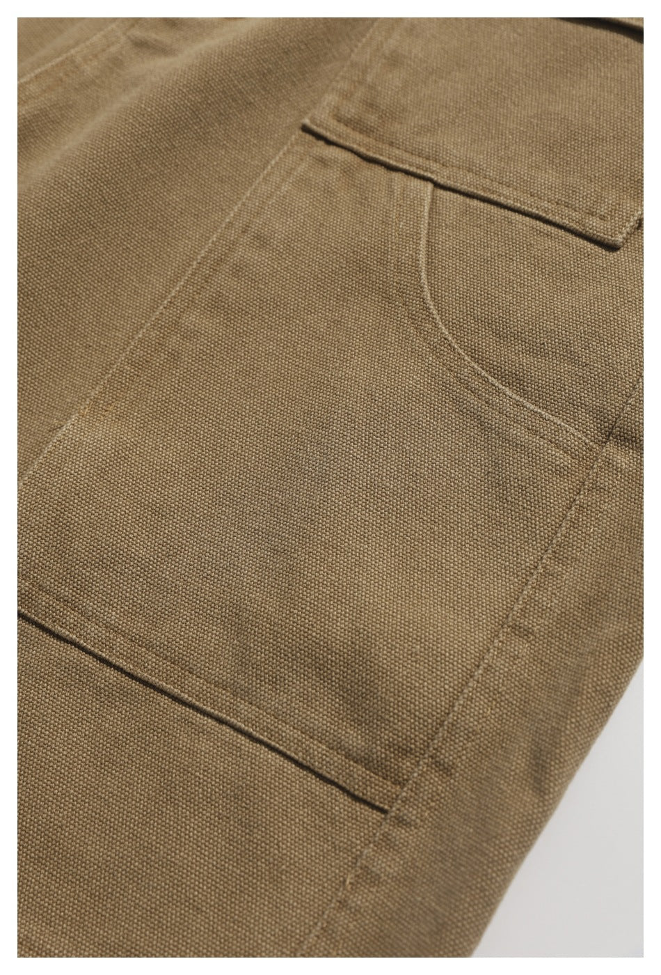 Detail of tan utility trousers showing a curved pocket opening, patch-pocket edge, and stitched seams.