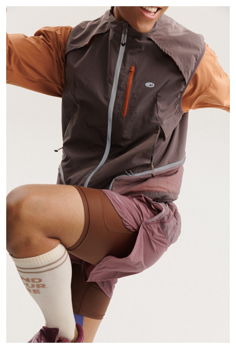 Close-up of a model wearing a dark taupe sleeveless zip vest over a rust-colored top and dusty rose shorts.