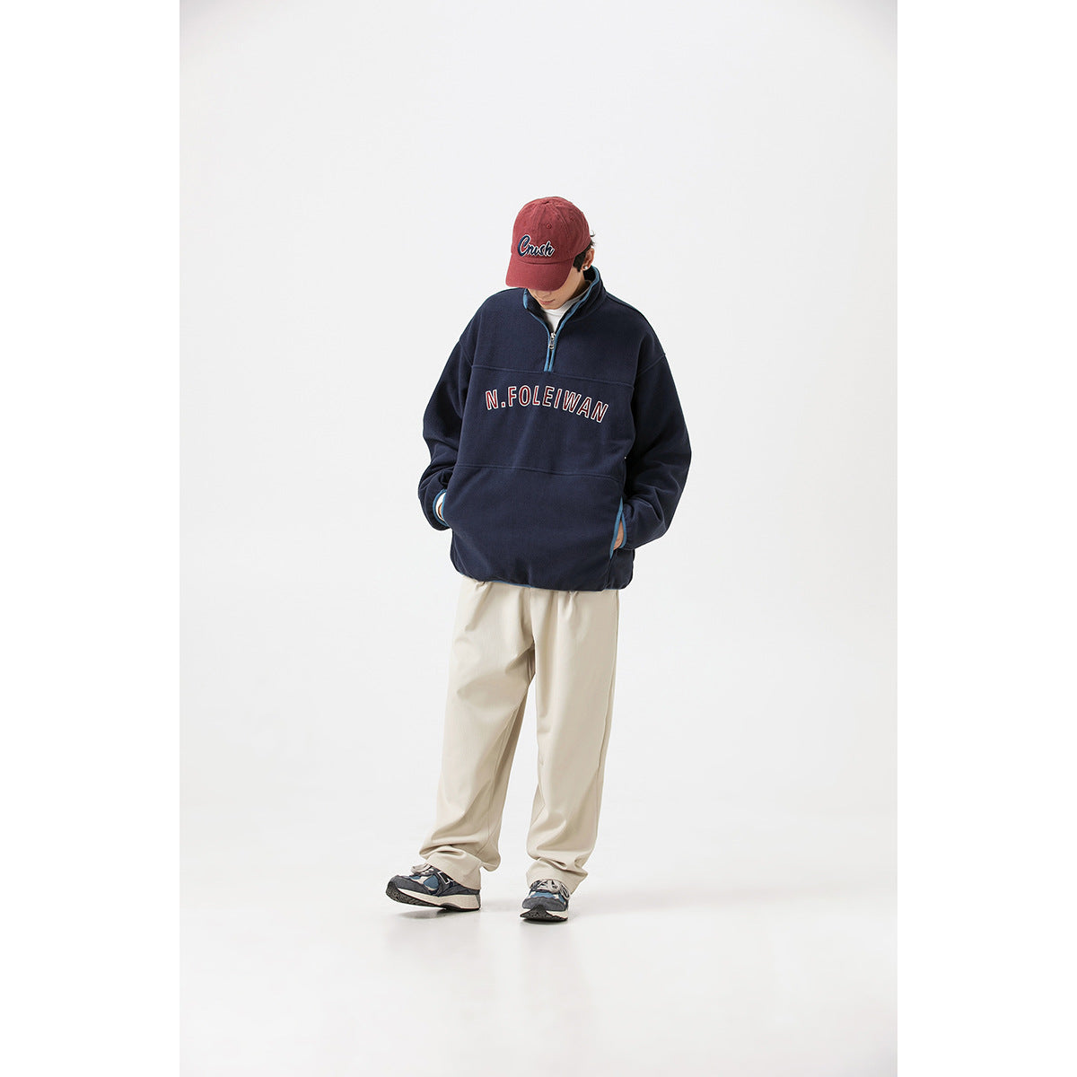 Model wearing a navy quarter-zip pullover with blue trim and chest lettering, cream trousers, and a red cap.