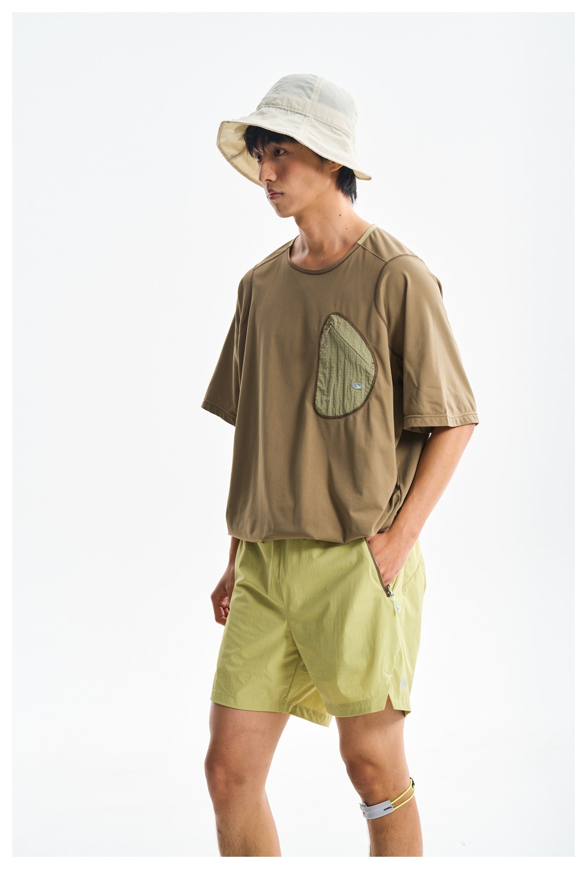 Angled view of a model wearing a brown T-shirt with a contrasting chest pocket and light yellow-green shorts.