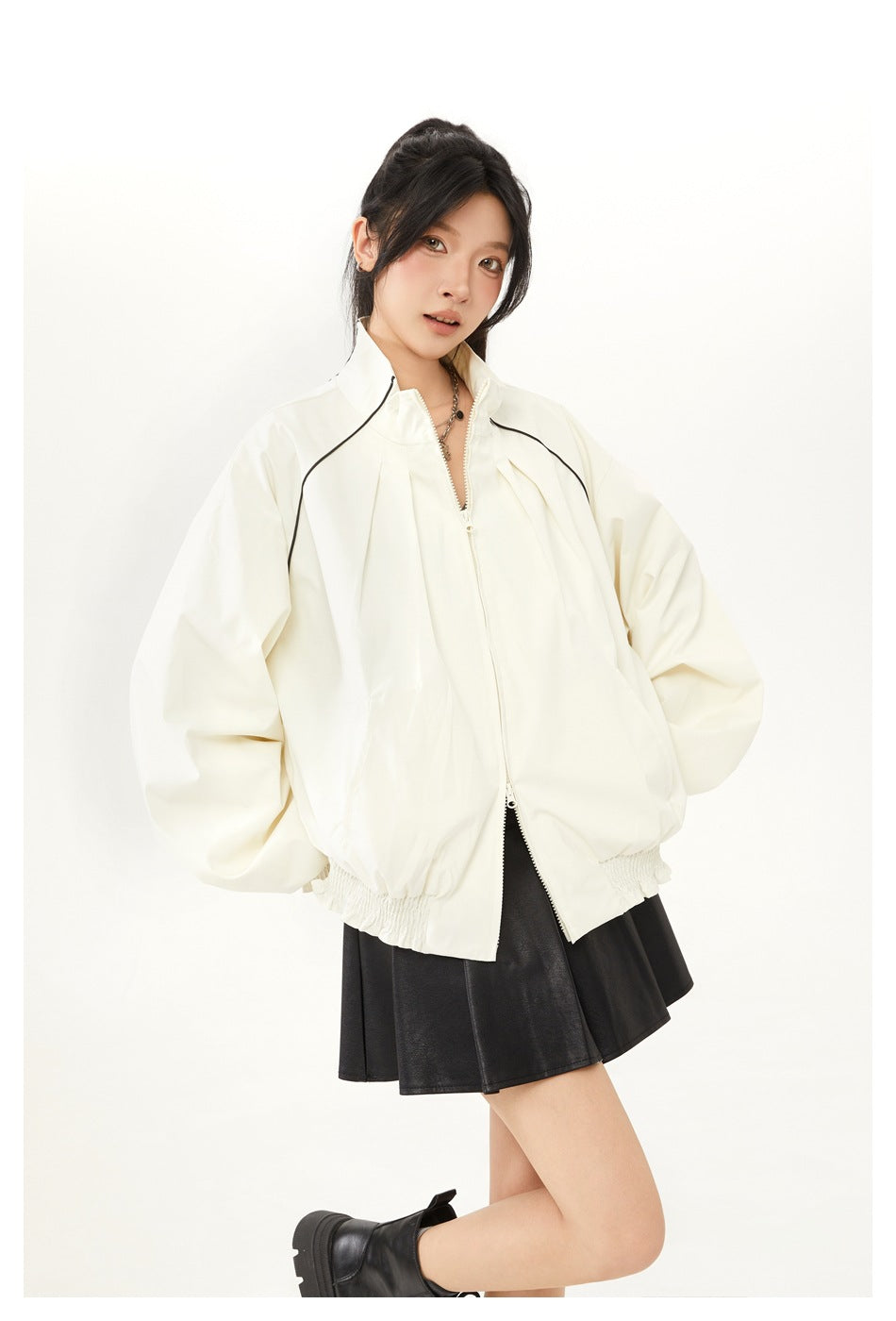 Model wearing an ivory oversized jacket with black piping and a black flared skirt, shown with one leg bent.