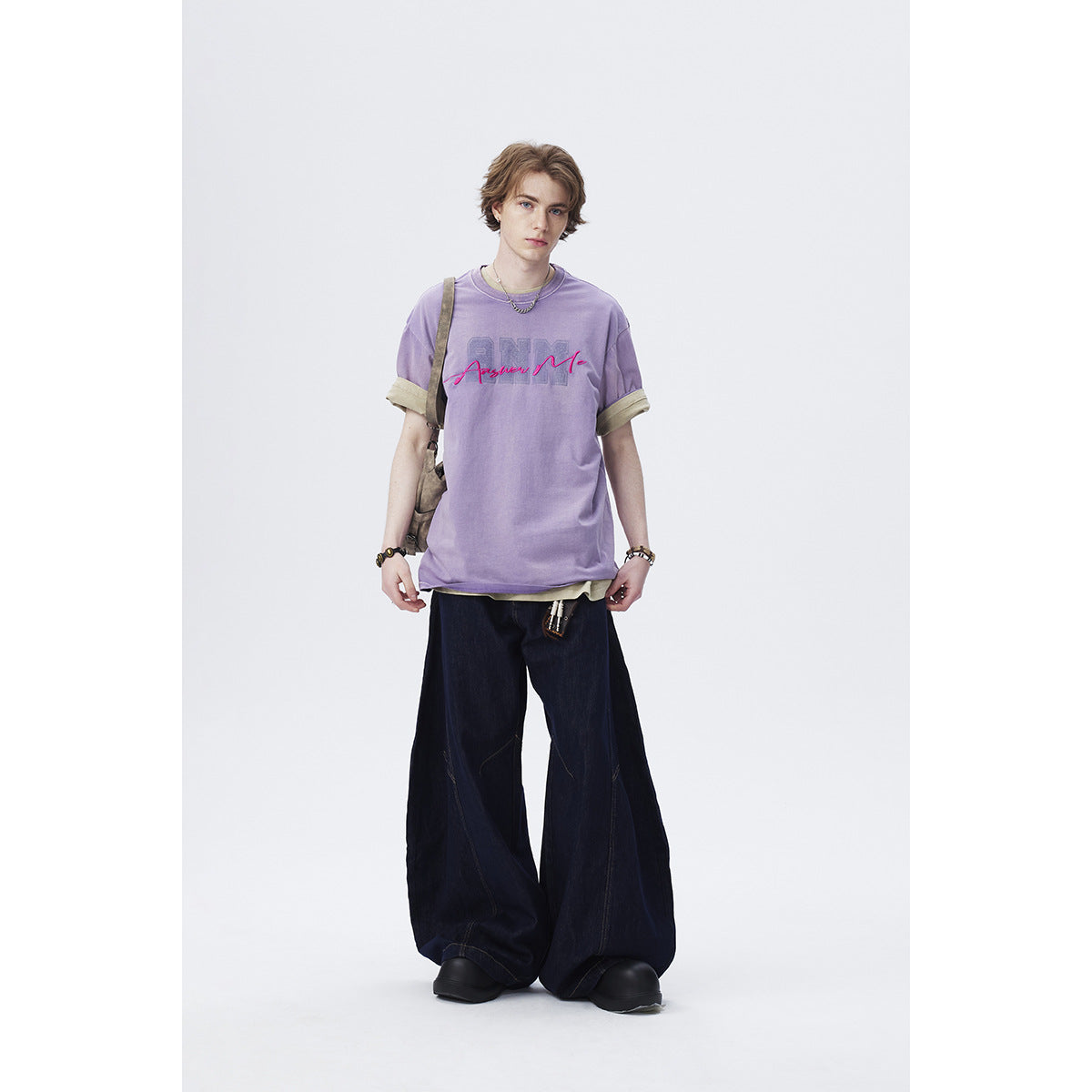 Model wearing a lavender graphic T-shirt over a light shirt, with very wide dark jeans and black shoes.