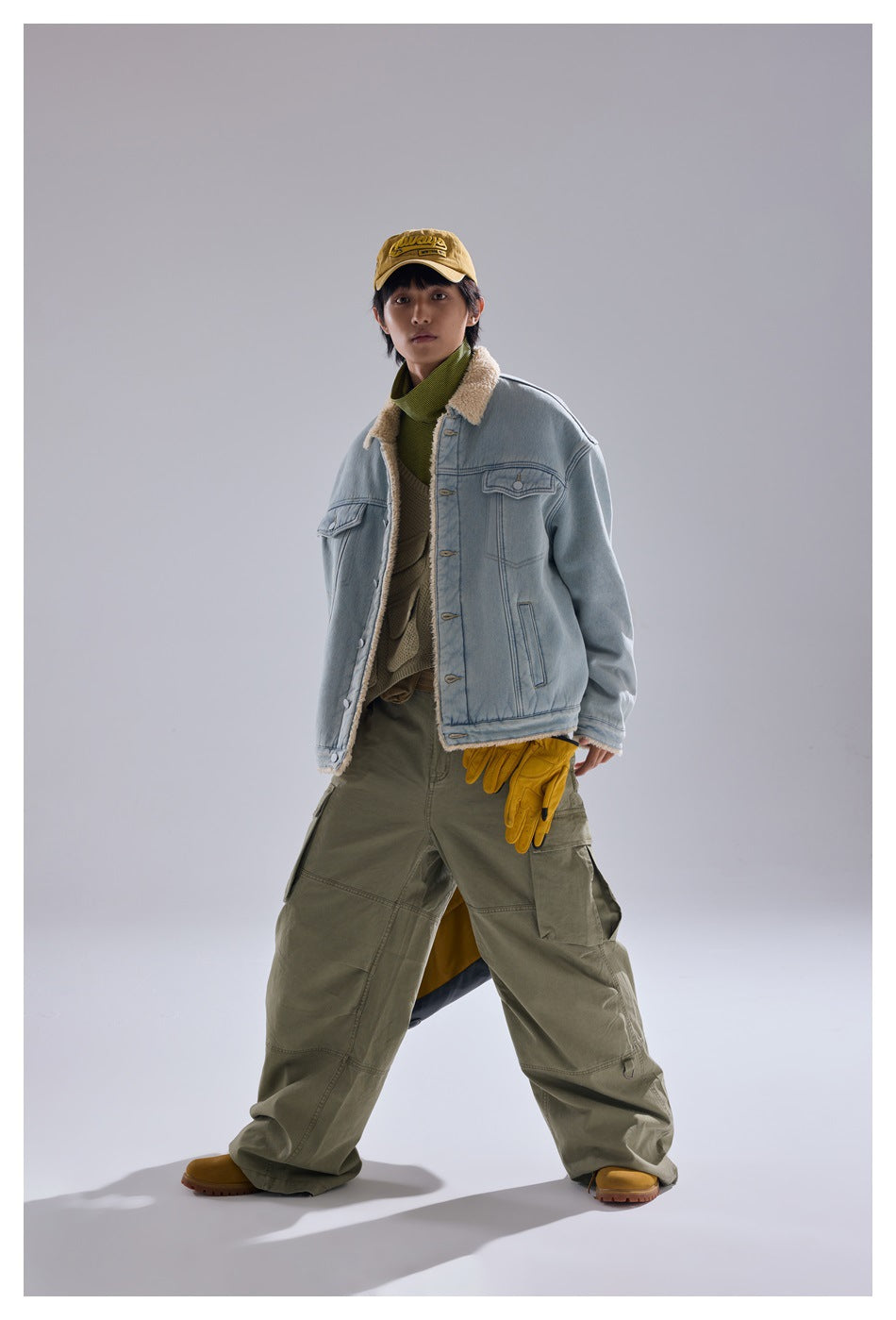 Model wearing a light blue sherpa-collar denim jacket with olive cargo pants and a yellow cap.