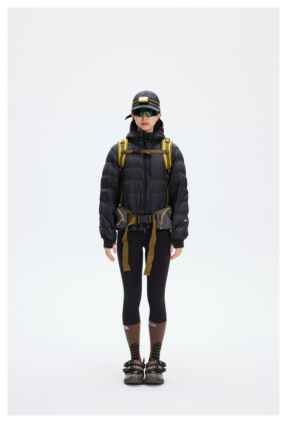 Front view of a model wearing a black hooded puffer jacket with a hiking backpack and waist belt.