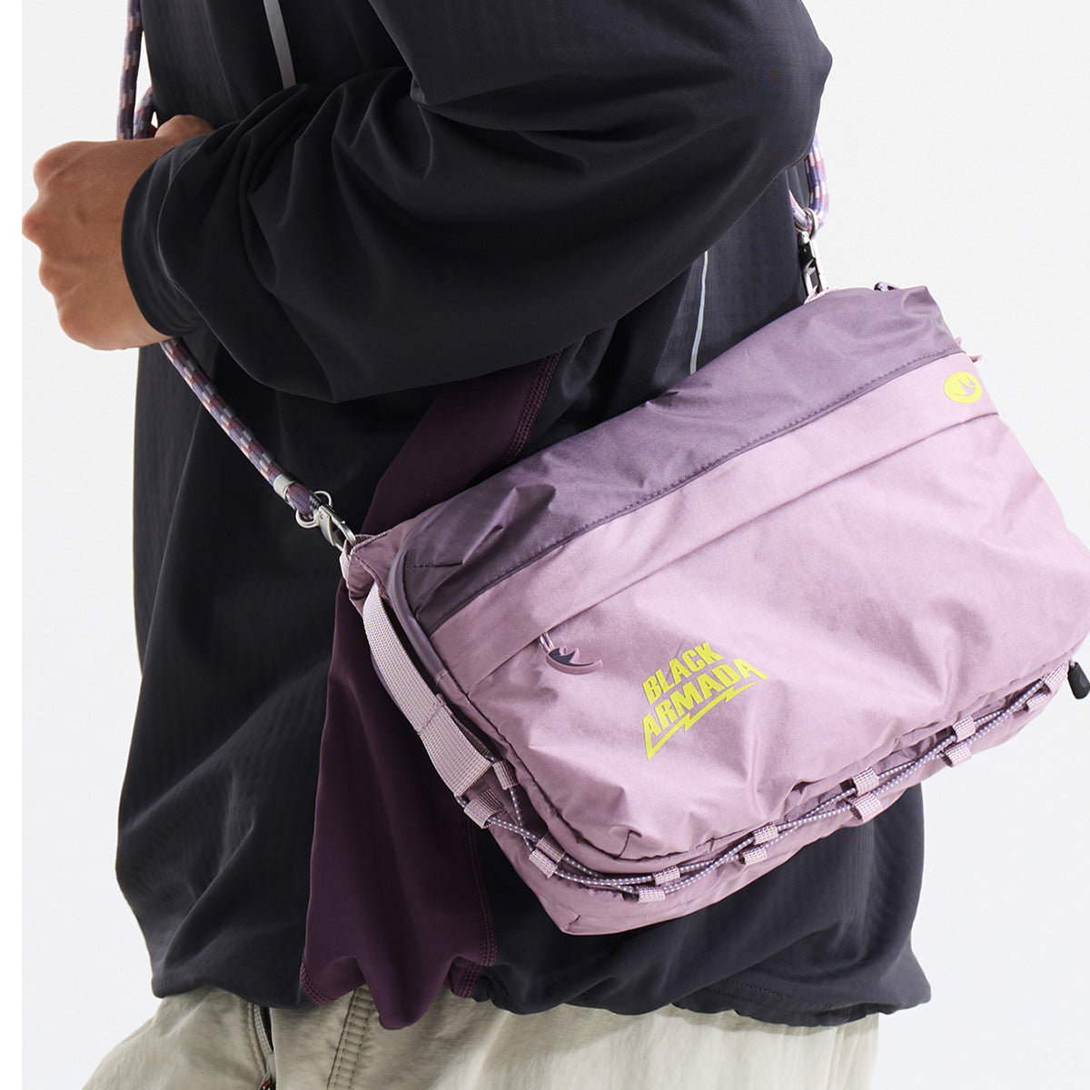 A person wears a lilac crossbody utility bag with a wide flap, cord details and yellow Black Armada lettering.