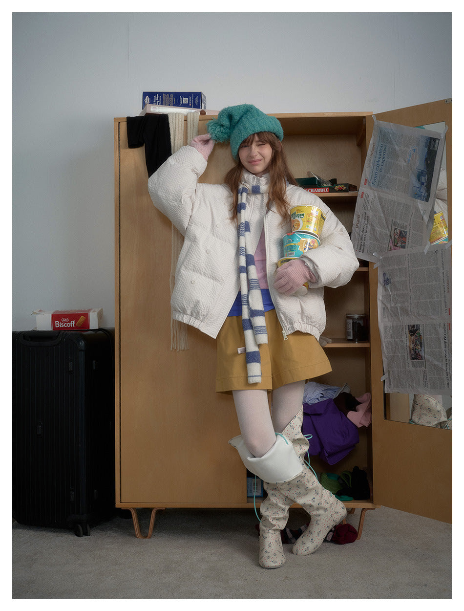 Model in white puffer jacket and striped scarf with one hand raised to a teal beanie, standing by a wooden wardrobe