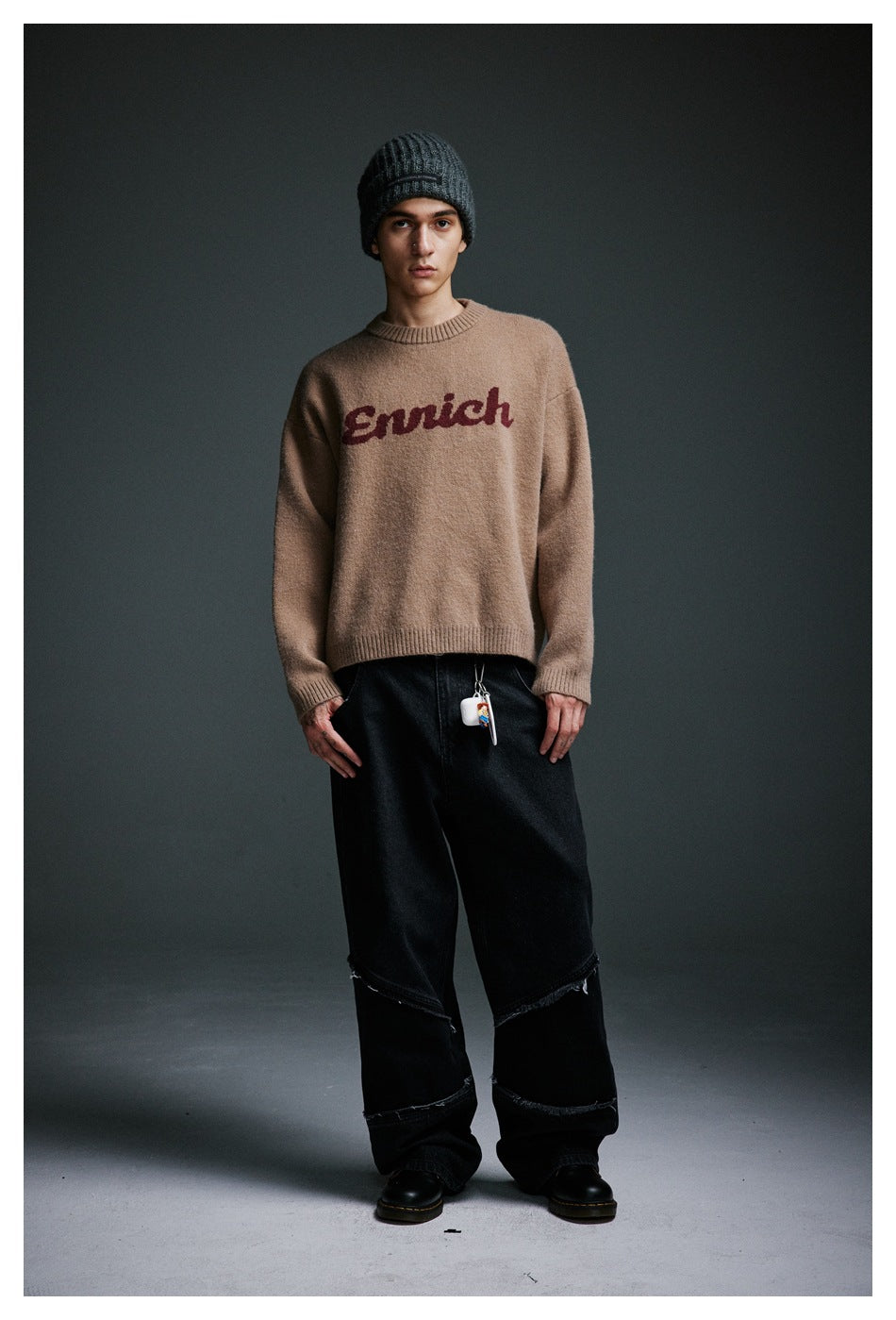 Full-length front view of a model wearing a tan sweater with burgundy script lettering, loose black jeans, and a dark beanie.