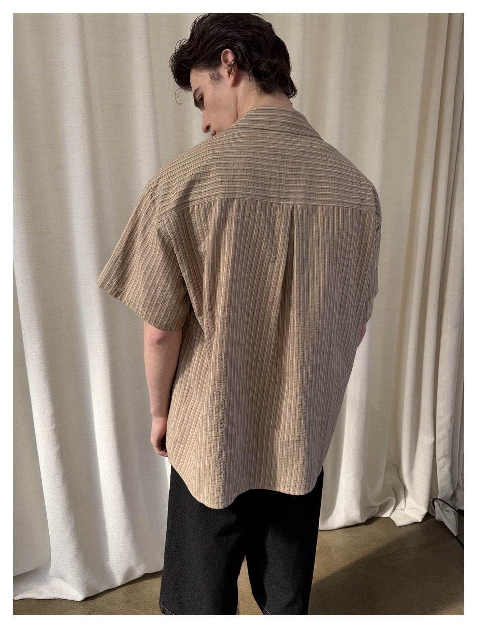 Rear view of a model wearing a taupe striped short-sleeve shirt with a pleated back yoke.