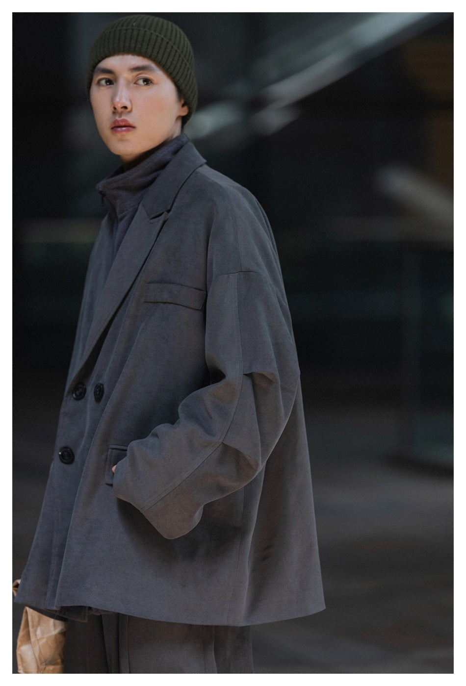 Model in a slate-gray oversized coat and olive ribbed beanie, shown from a three-quarter angle.