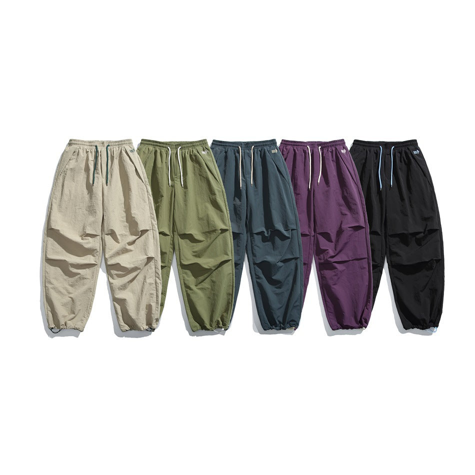 Five pairs of loose drawstring pants shown together in beige, olive, dark teal, purple, and black.