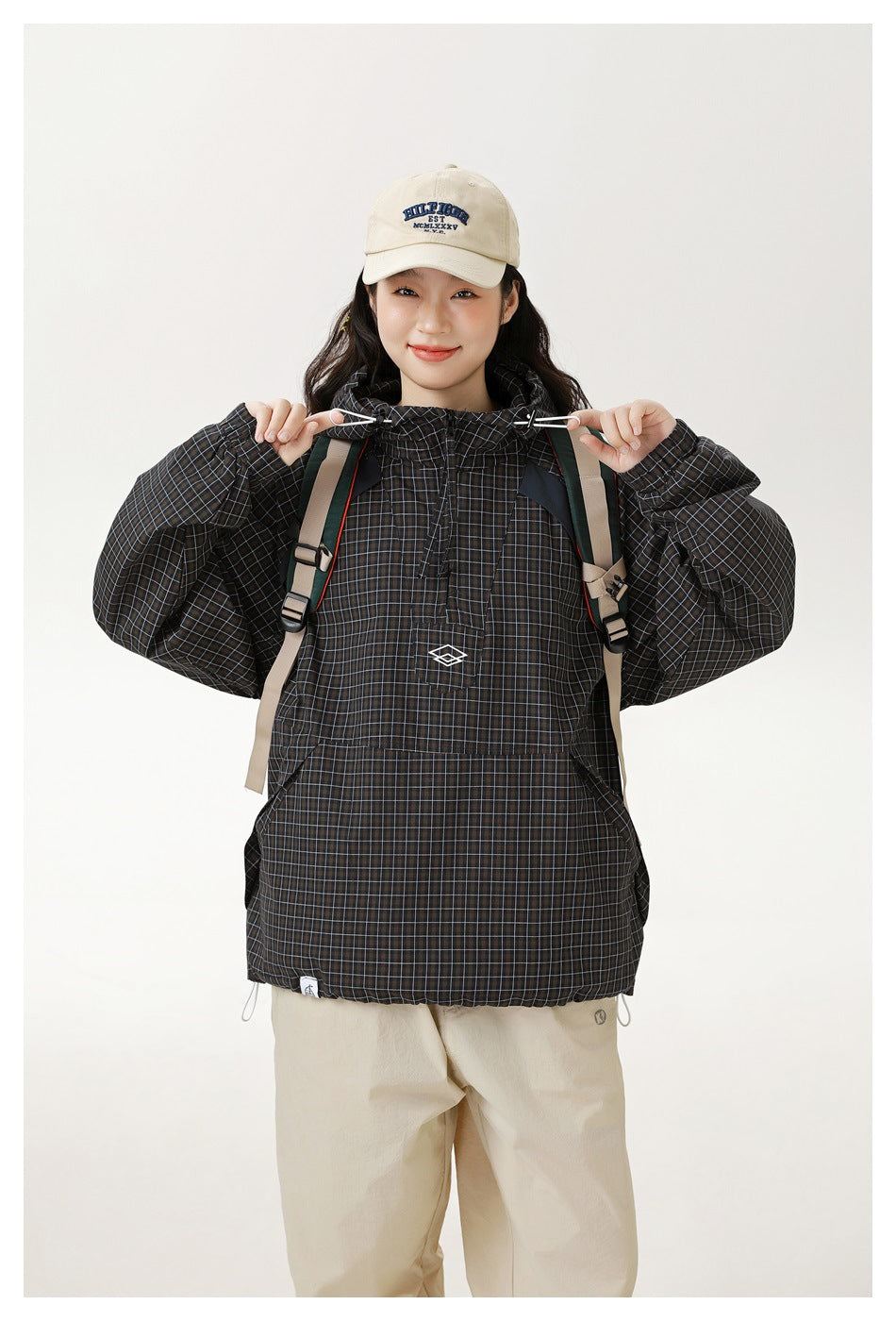 Model in the grid check hooded jacket holding the hood drawstring toggles, wearing a backpack and cream cap, close view