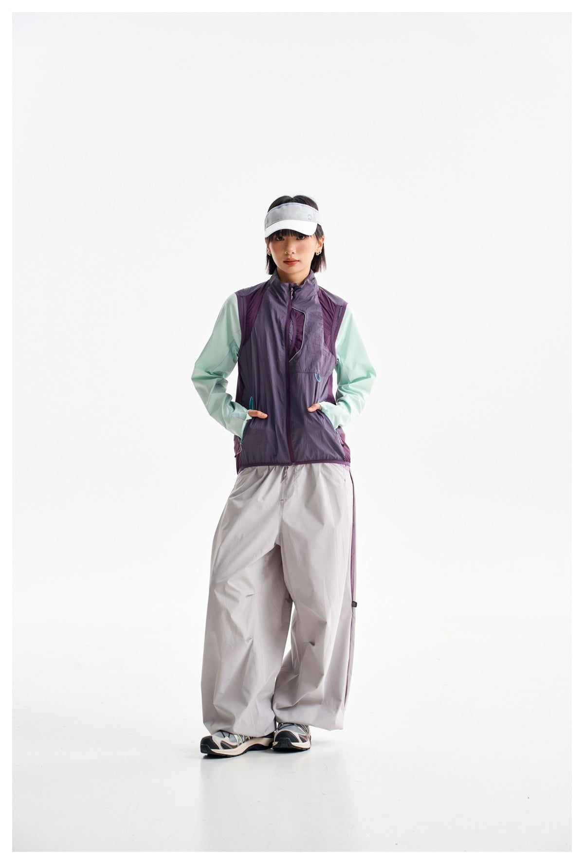 Full-length front view of a woman wearing a plum utility vest over a mint long-sleeve top with loose gray pants and a white