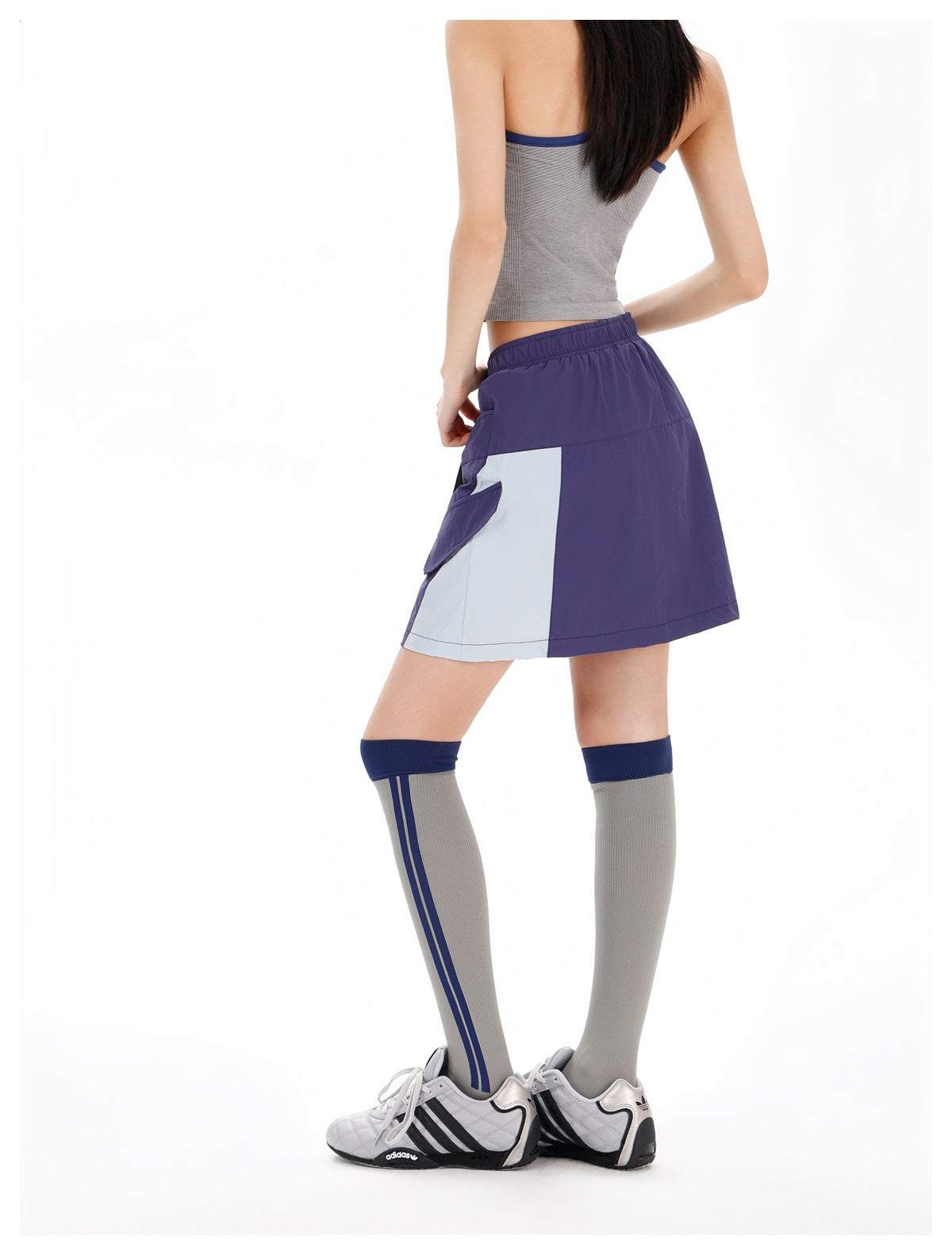 Rear three-quarter view of a model wearing a purple mini skirt with a pale gray side panel and flap pocket.