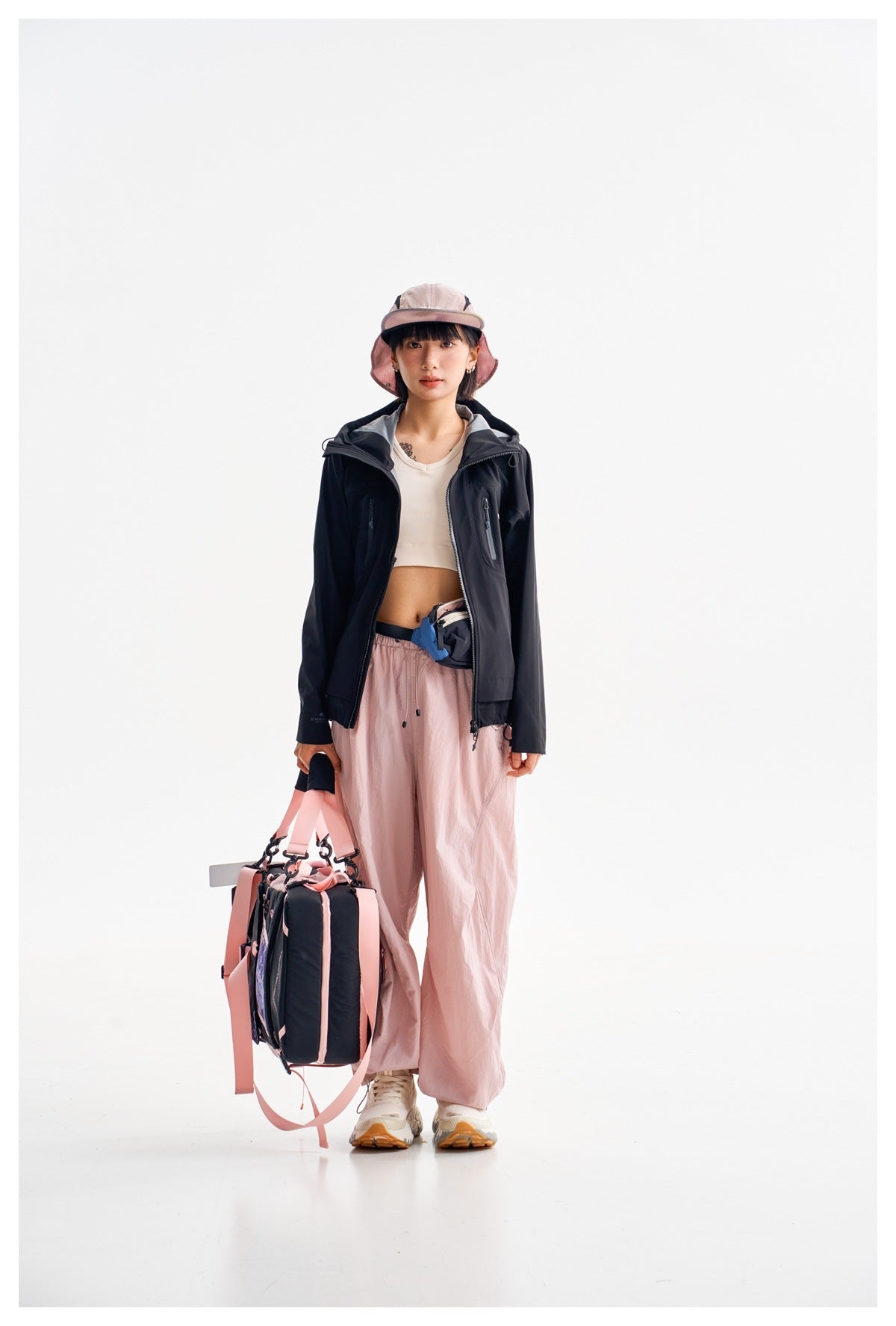 Model wearing a navy zip jacket with pink trousers and a pink sun hat, carrying a large bag.