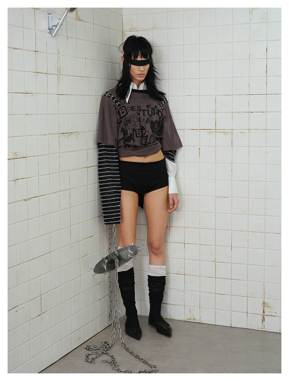 Model leaning in a tiled corner wearing the gray graffiti top with striped sleeves, blindfold and black shorts