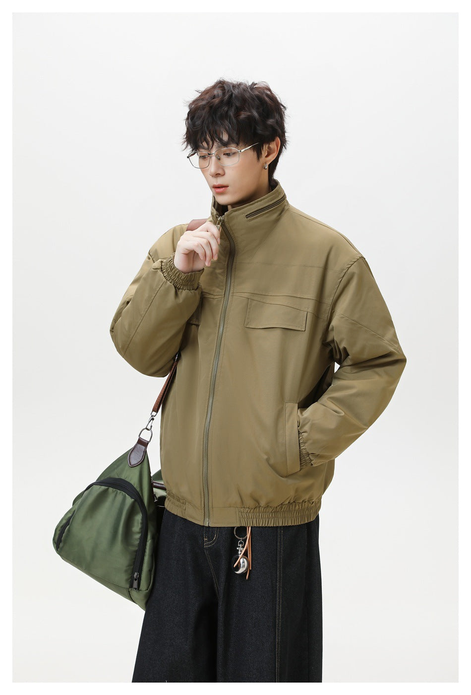 Model wearing a zipped olive-brown jacket with a stand collar and flap chest pocket, carrying a green bag.