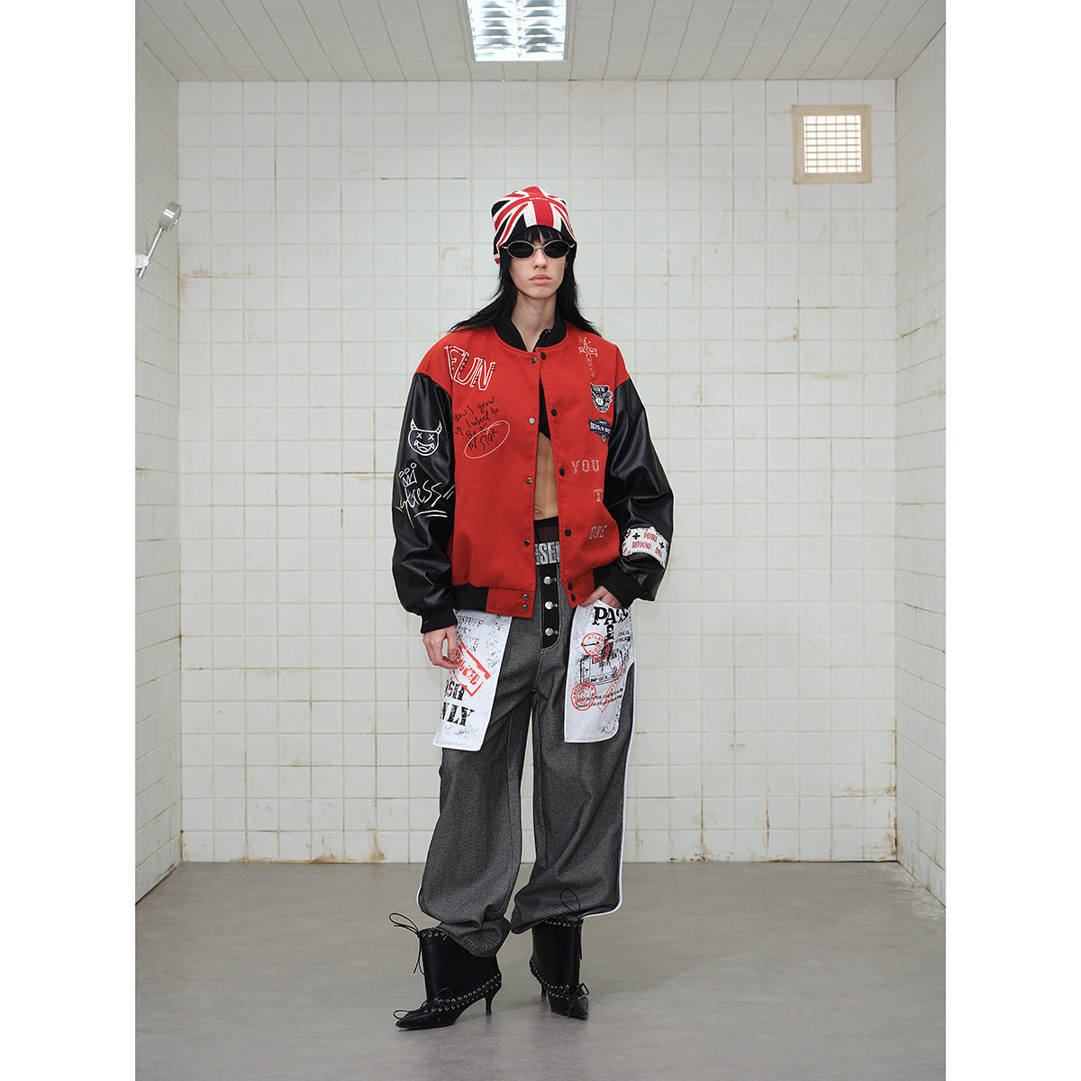 Full-length model wearing a red-and-black graphic varsity jacket, gray trousers, black boots, and a Union Jack beanie.