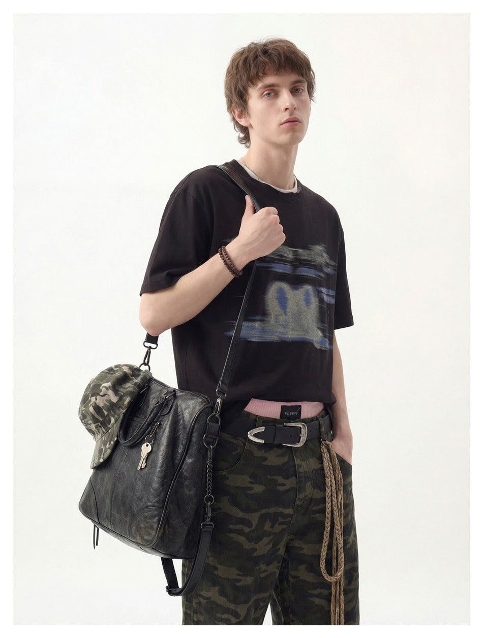 Model wearing a black graphic T-shirt with loose camouflage trousers and carrying a black bag.