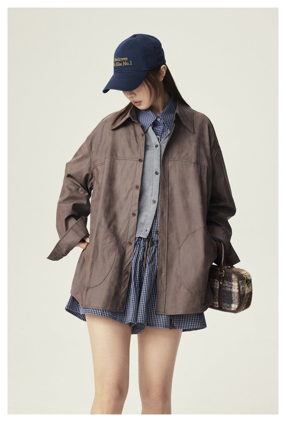 Model wearing a brown oversized button-front shirt jacket with curved pockets over a gray vest and plaid shirt