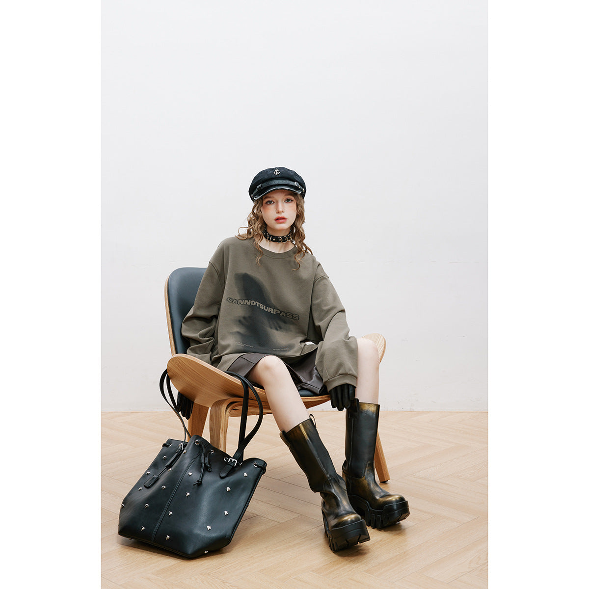 Model seated in an oversized olive-gray graphic sweatshirt, with a black studded tote and boots.