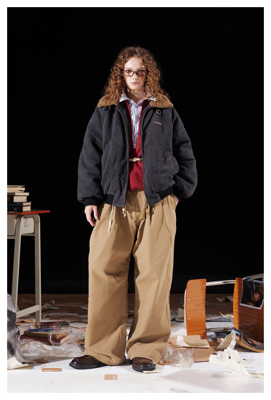 Full-length model wearing a charcoal jacket with a tan collar, red sweater, striped shirt and loose tan trousers.