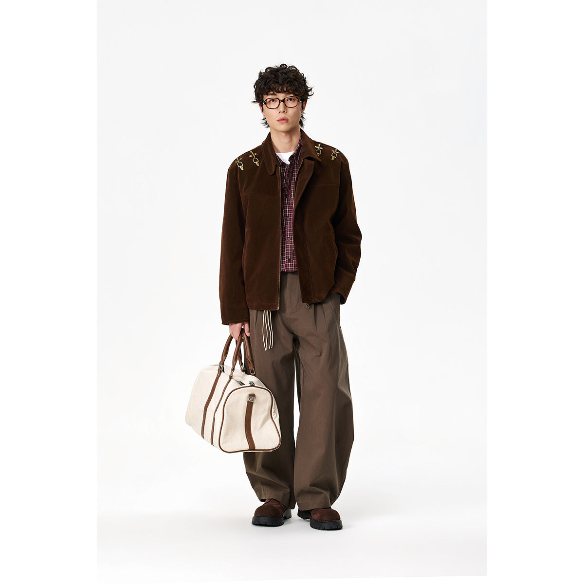 Full-length model wearing a dark brown jacket with gold shoulder hardware, plaid shirt, taupe trousers, and carrying a cream