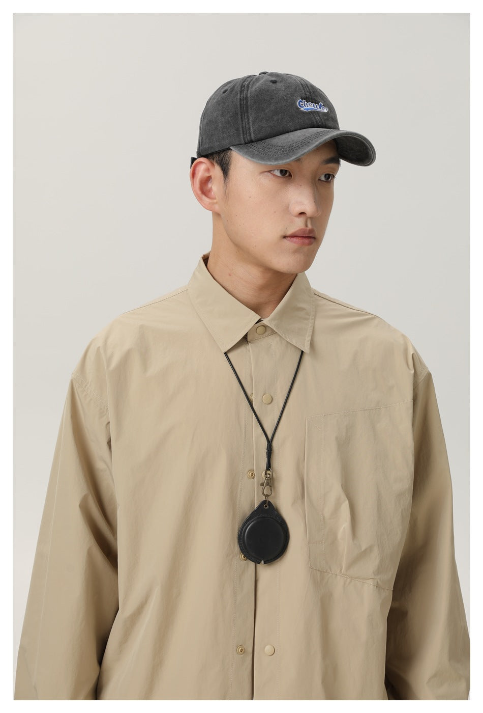 Model wearing a beige button-front shirt with a chest pocket, black lanyard, and charcoal cap with embroidered lettering.