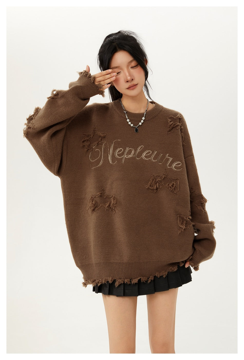 Close view of a model in a brown oversized distressed sweater with embroidered script and a black pleated mini skirt.
