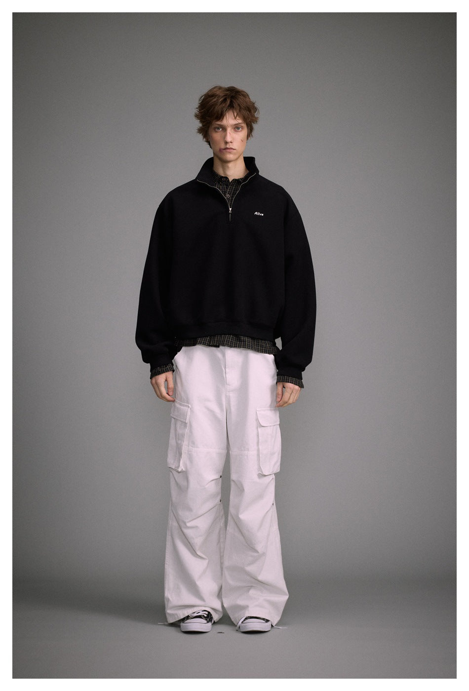 Model wearing black quarter-zip sweatshirt over a plaid shirt with white cargo pants, front view