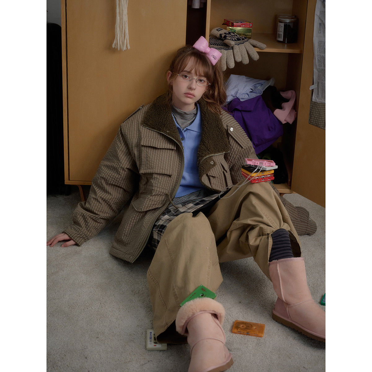 Seated model wearing an open taupe checked jacket with a dark fleece collar and large flap pockets.