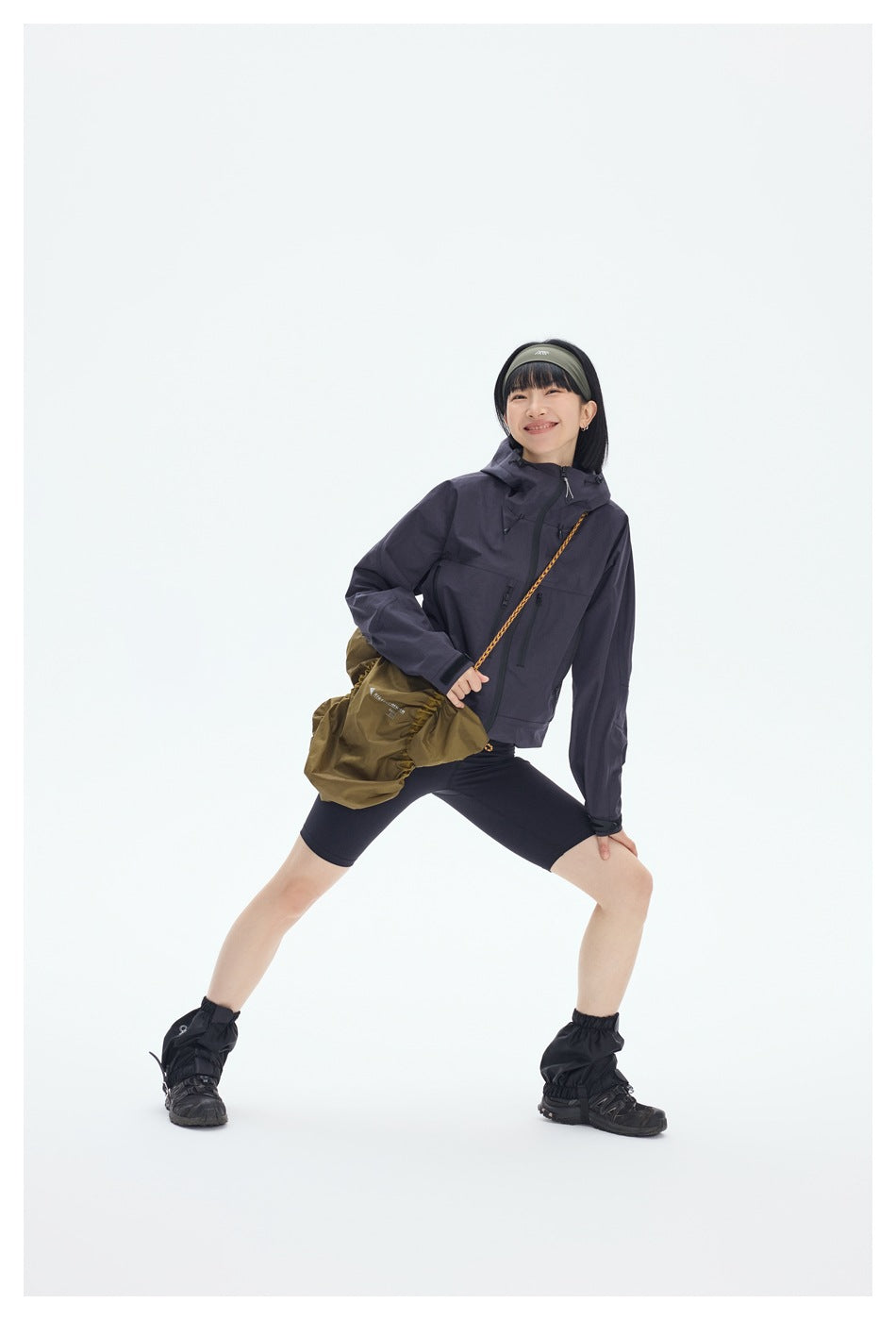 A model poses in a charcoal hooded jacket with front zip pockets, black shorts, and a crossbody bag.