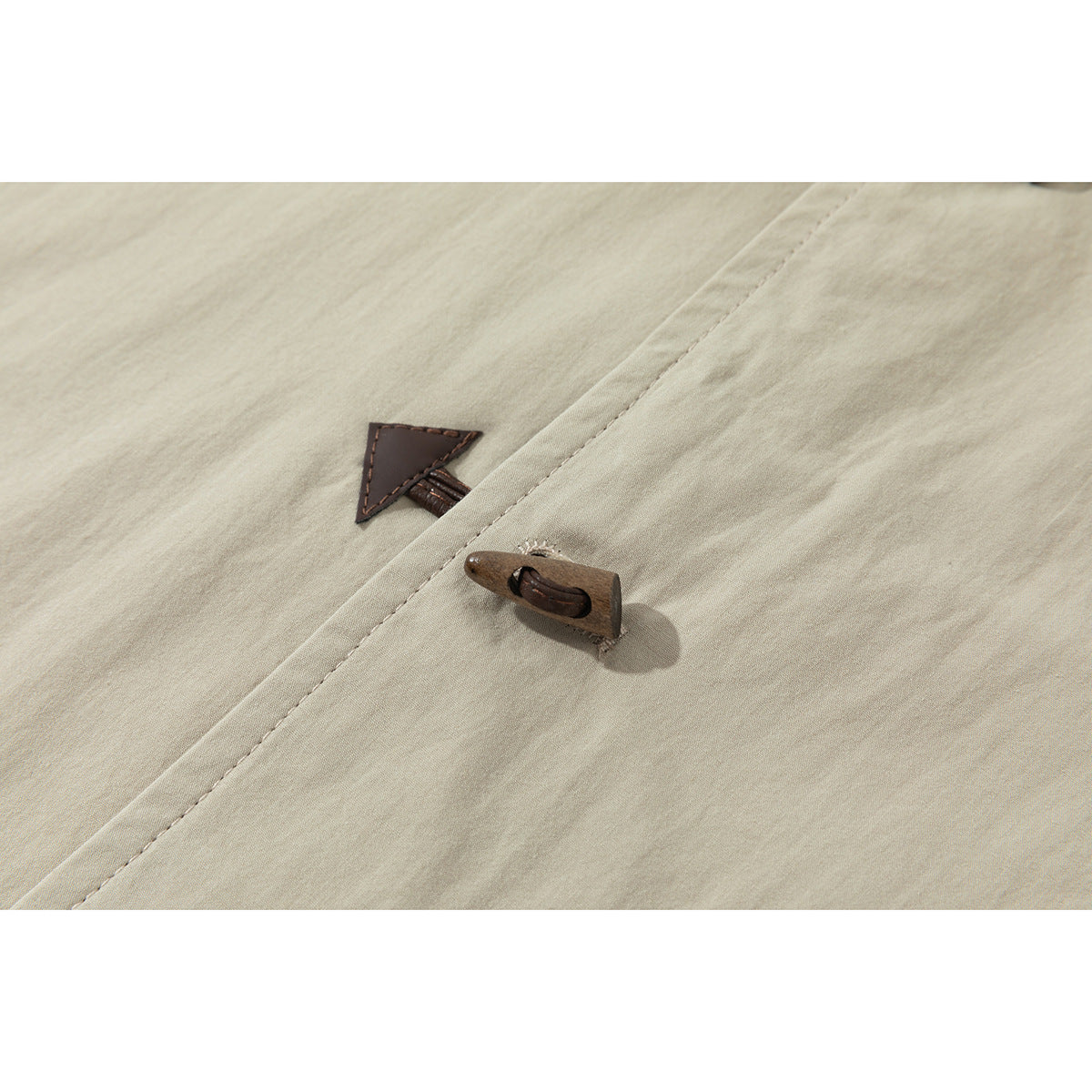 Close-up of a beige jacket's brown toggle closure and triangular leather-like tab on the front placket.