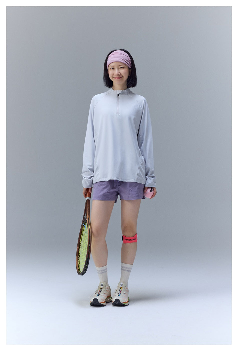 Full-length model wearing a pale gray quarter-zip top and lavender shorts with a tennis racket.